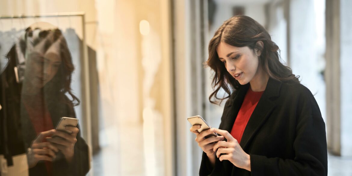 Woman wearing red top and black coat on her phone in front of store window