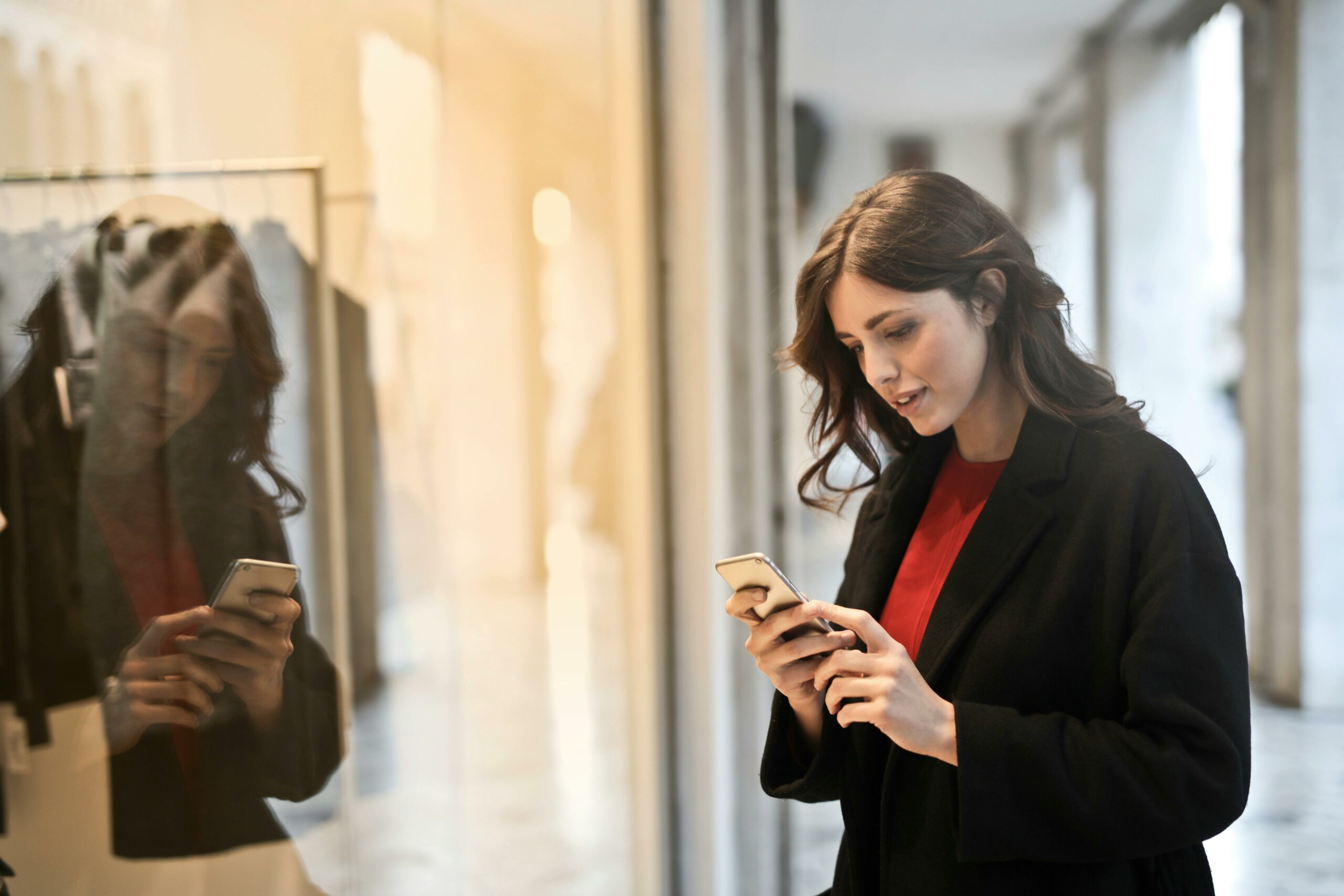 Woman wearing red top and black coat on her phone in front of store window